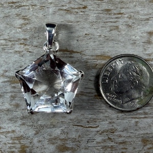 May include: A clear, faceted star-shaped gemstone pendant set in a silver-toned metal bail. The pendant is positioned next to a United States dime for scale, which measures approximately 1.79 cm in diameter, providing a clear size reference.