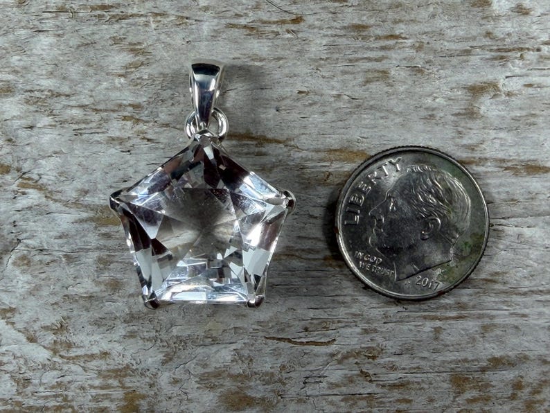 May include: A clear, faceted star-shaped gemstone pendant set in a silver-toned metal bail. The pendant is positioned next to a United States dime for scale, which measures approximately 1.79 cm in diameter, providing a clear size reference.