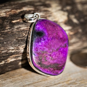 May include: A pendant with a large, polished, purple stone set in a silver-colored metal frame. The stone has a mottled appearance with darker purple and black inclusions. The pendant is displayed on a wooden surface.