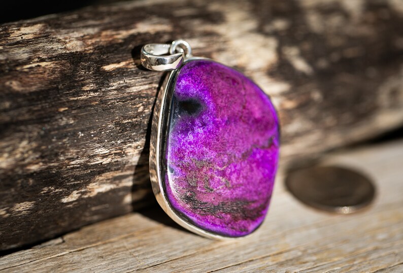 May include: A pendant with a large, polished, purple stone set in a silver-colored metal frame. The stone has a mottled appearance with darker purple and black inclusions. The pendant is displayed on a wooden surface.