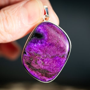 May include: A vibrant purple pendant with a silver-colored bezel. The stone has a marbled appearance with dark purple and black veining. The pendant is held up by a silver bail.