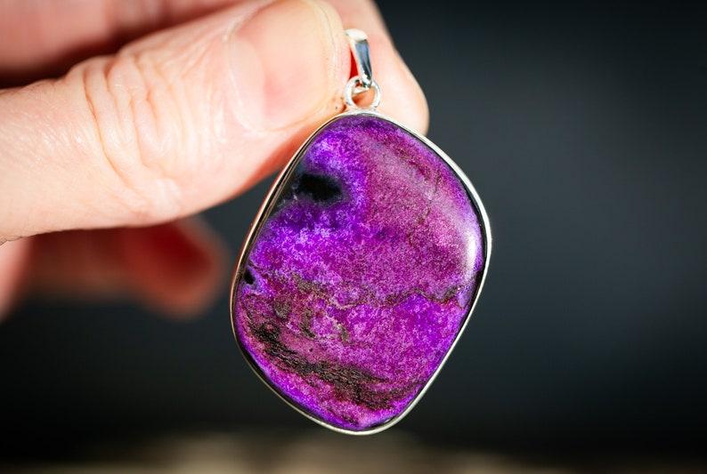 May include: A vibrant purple pendant with a silver-colored bezel. The stone has a marbled appearance with dark purple and black veining. The pendant is held up by a silver bail.