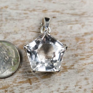 May include: A clear, faceted, star-shaped pendant with a silver-toned metal bail at the top. The pendant is positioned next to a United States dime for scale, showing the relative size of the jewellery piece against the coin.