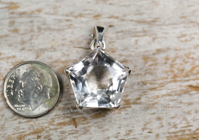 May include: A clear, faceted, star-shaped pendant with a silver-toned metal bail at the top. The pendant is positioned next to a United States dime for scale, showing the relative size of the jewellery piece against the coin.