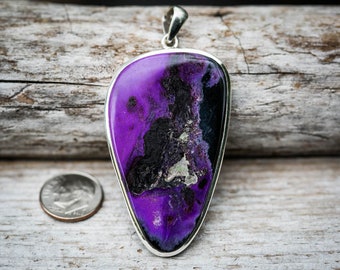 Puede incluir: Un colgante en forma de lágrima con una piedra preciosa púrpura y negra en un engaste de plata. El colgante tiene un patrón único y arremolinado.