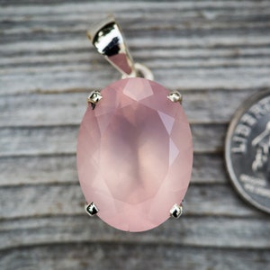 May include: A pale pink oval-shaped rose quartz pendant with silver accents. The pendant is set in a silver bezel and has a faceted design. The pendant is approximately the size of a US dime.