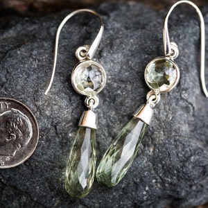 May include: A pair of silver dangle earrings with teardrop-shaped green gemstones. The earrings have a round gemstone at the top and a teardrop-shaped gemstone hanging below.