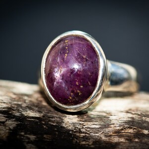 May include: A silver ring featuring a large, oval-shaped, deep purple gemstone with gold flecks. The ring is set in a polished silver band, showcasing the stone's rich color and texture. The ring is displayed on a piece of wood.