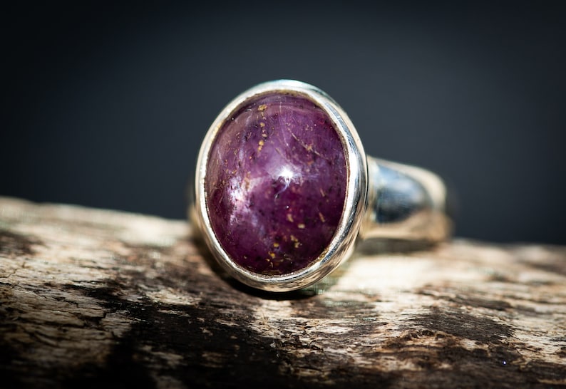 May include: A silver ring featuring a large, oval-shaped, deep purple gemstone with gold flecks. The ring is set in a polished silver band, showcasing the stone's rich color and texture. The ring is displayed on a piece of wood.