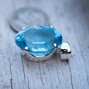 Pu&ograve; includere: Un ciondolo d'argento con una grande pietra ovale di colore blu chiaro. Il ciondolo &egrave; su una superficie di legno bianco.