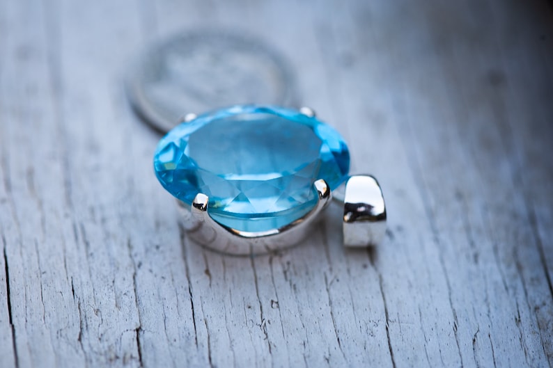 Pu&ograve; includere: Un ciondolo d'argento con una grande pietra ovale di colore blu chiaro. Il ciondolo &egrave; su una superficie di legno bianco.