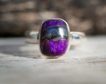 May include: A silver ring featuring a rectangular, polished stone with a vibrant purple hue, accented by bands of gray and black. The ring's setting is a simple, polished silver band, showcasing the stone's unique color and pattern.