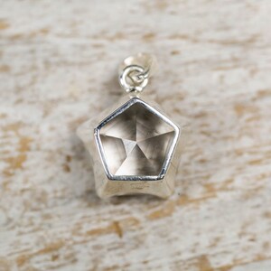 May include: A silver-toned pendant featuring a pentagonal shape with a faceted, translucent, light brown gemstone set in the centre. The pendant includes a small silver-toned metal loop at the top for attachment to a chain or cord.