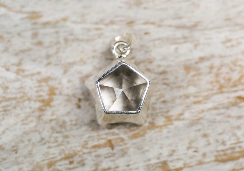 May include: A silver-toned pendant featuring a pentagonal shape with a faceted, translucent, light brown gemstone set in the centre. The pendant includes a small silver-toned metal loop at the top for attachment to a chain or cord.