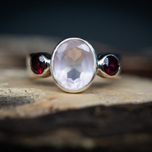 May include: A silver ring with a large oval-shaped rose quartz gemstone and two smaller round garnet gemstones on either side.