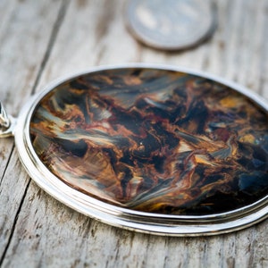 May include: Oval pendant with a silver-colored setting. The pendant features a polished stone with a swirling pattern of brown, blue, and gold colors. The pendant is displayed on a wooden surface.