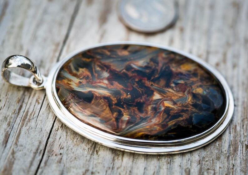 May include: Oval pendant with a silver-colored setting. The pendant features a polished stone with a swirling pattern of brown, blue, and gold colors. The pendant is displayed on a wooden surface.