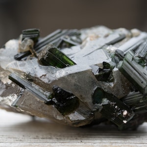 May include: A close-up of a raw green tourmaline crystal cluster. The dark green, elongated crystals are set in a matrix of clear quartz and other minerals. The natural formation has a rough, textured appearance.