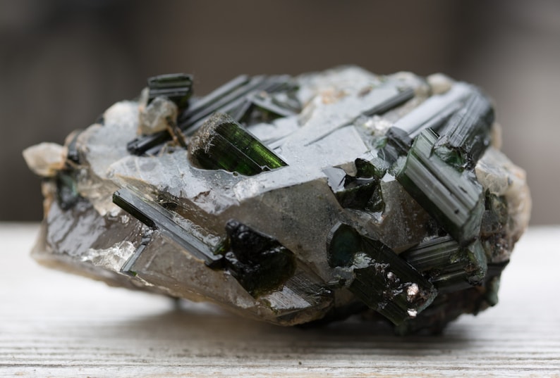 May include: A close-up of a raw green tourmaline crystal cluster. The dark green, elongated crystals are set in a matrix of clear quartz and other minerals. The natural formation has a rough, textured appearance.