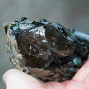 May include: A raw gemstone with a mix of dark brown, clear, and teal crystals. The mineral specimen has a rugged, natural appearance, with visible crystalline structures and a textured surface. The overall colour palette is earthy and natural.