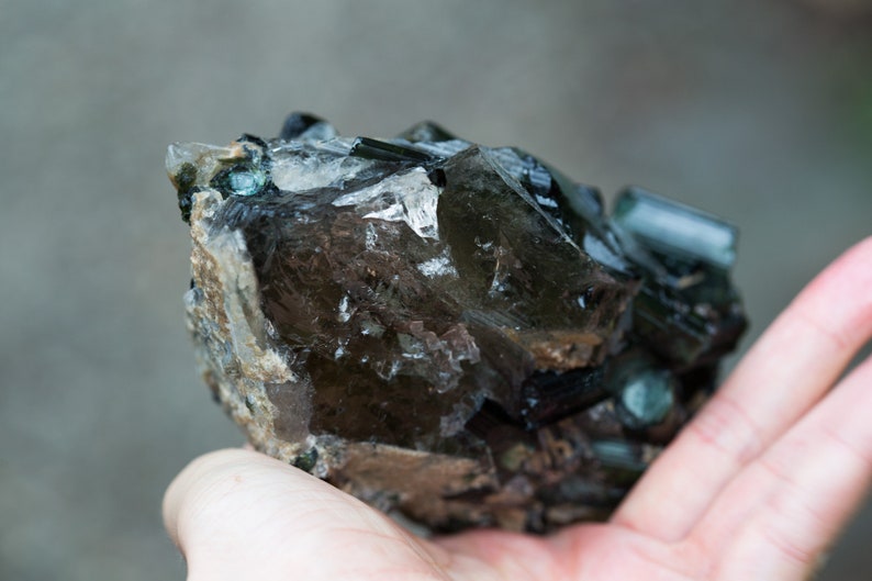 May include: A raw gemstone with a mix of dark brown, clear, and teal crystals. The mineral specimen has a rugged, natural appearance, with visible crystalline structures and a textured surface. The overall colour palette is earthy and natural.