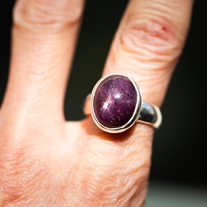 May include: A sterling silver ring featuring a large, oval-shaped, deep purple gemstone with gold flecks. The gemstone is set in a simple bezel setting, creating a classic and elegant design. The ring is shown on a finger.