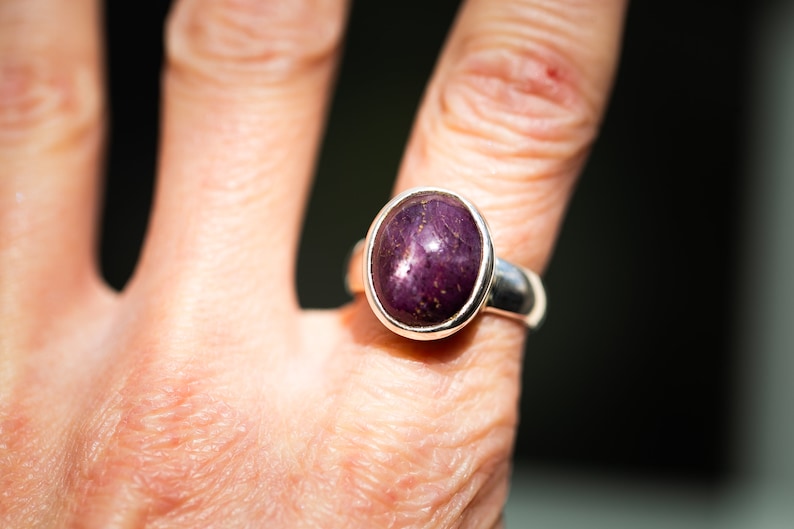 May include: A sterling silver ring featuring a large, oval-shaped, deep purple gemstone with gold flecks. The gemstone is set in a simple bezel setting, creating a classic and elegant design. The ring is shown on a finger.