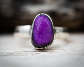 May include: A close-up of a silver ring featuring a large, oval-shaped purple stone. The stone is set in a silver bezel and has a smooth, polished surface. The ring is set against a blurred wooden background.