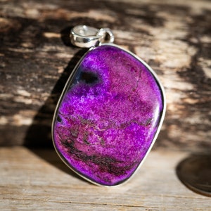 May include: A vibrant purple pendant with a silver bail. The gemstone has a unique, irregular shape and a textured surface with dark veins. The pendant is set against a wooden background, with a coin visible for scale.