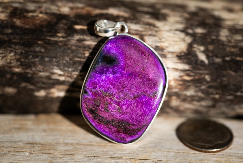 May include: A vibrant purple pendant with a silver bail. The gemstone has a unique, irregular shape and a textured surface with dark veins. The pendant is set against a wooden background, with a coin visible for scale.