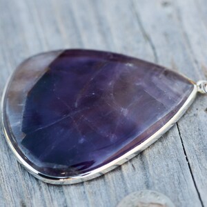 May include: A large, teardrop-shaped pendant with a polished, deep purple gemstone. The stone is set in a silver bezel and has a small loop for a chain. The pendant is displayed on a weathered wooden surface.