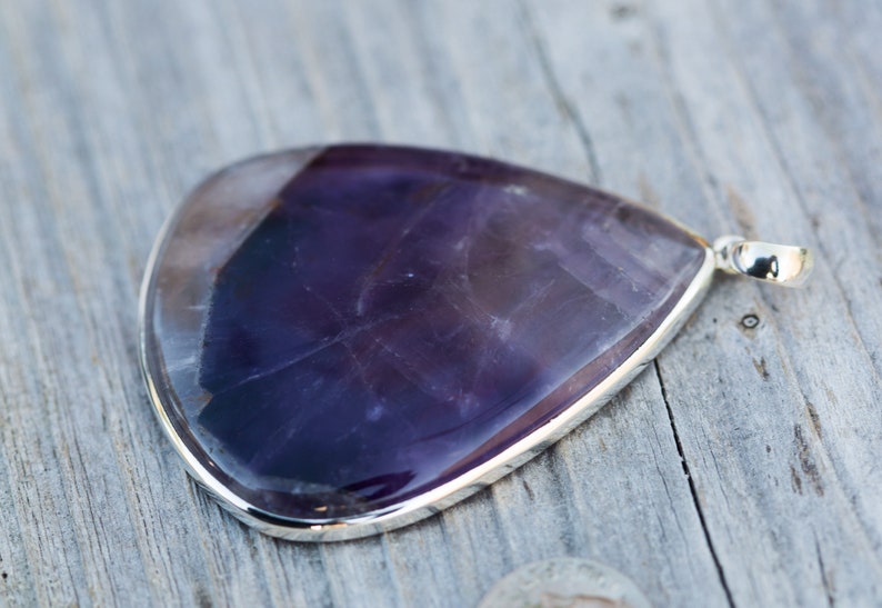 May include: A large, teardrop-shaped pendant with a polished, deep purple gemstone. The stone is set in a silver bezel and has a small loop for a chain. The pendant is displayed on a weathered wooden surface.