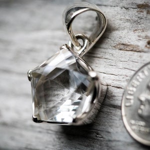 May include: A pentagonal faceted clear gemstone pendant set in a polished silver metal prong setting with a silver bail at the top. A United States dime is positioned to the right of the pendant to provide a visual scale reference for the size.