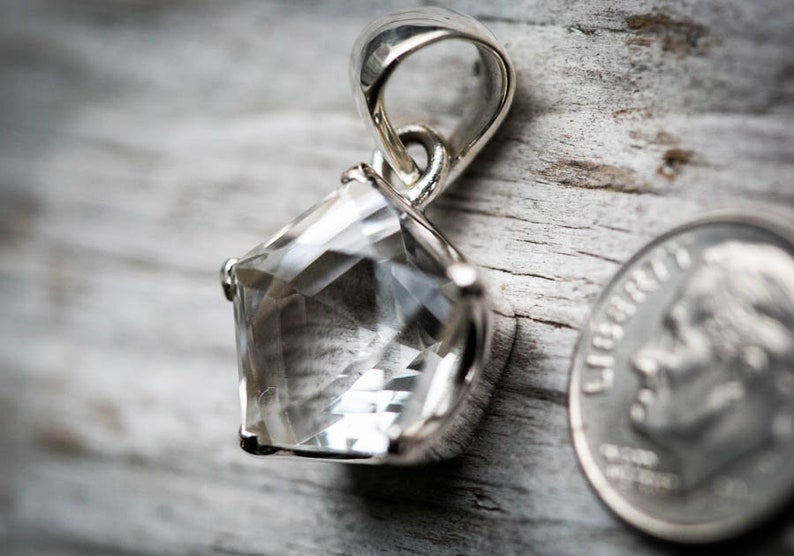 May include: A pentagonal faceted clear gemstone pendant set in a polished silver metal prong setting with a silver bail at the top. A United States dime is positioned to the right of the pendant to provide a visual scale reference for the size.