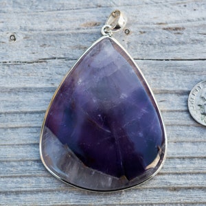 May include: Large, teardrop-shaped amethyst pendant with a silver-colored bezel and bail. The polished stone displays shades of purple and white. A U.S. dime is next to the pendant for size comparison. The pendant is on a weathered wooden surface.