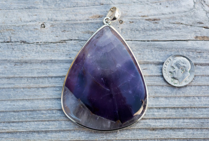 May include: Large, teardrop-shaped amethyst pendant with a silver-colored bezel and bail. The polished stone displays shades of purple and white. A U.S. dime is next to the pendant for size comparison. The pendant is on a weathered wooden surface.