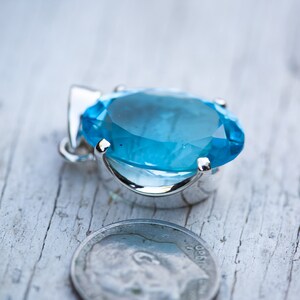 Pu&ograve; includere: Un ciondolo d'argento con una grande pietra ovale di colore blu chiaro. Il ciondolo &egrave; posto su una superficie di legno bianco accanto a una moneta da dieci centesimi di dollaro americano.
