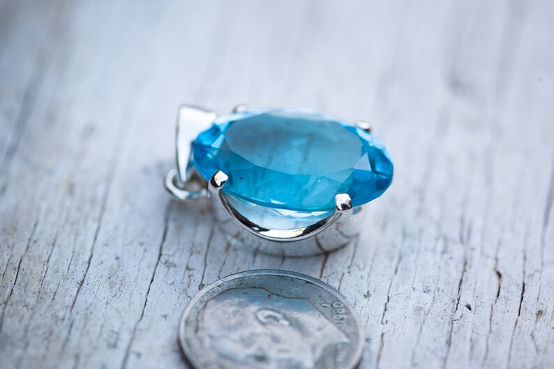 Pu&ograve; includere: Un ciondolo d'argento con una grande pietra ovale di colore blu chiaro. Il ciondolo &egrave; posto su una superficie di legno bianco accanto a una moneta da dieci centesimi di dollaro americano.