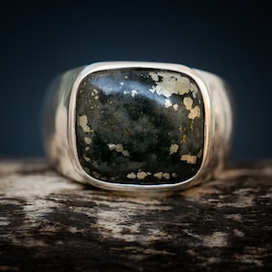 May include: A silver ring with a square, dark green gemstone with gold flecks. The ring is set on a wooden surface.