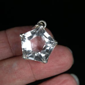 May include: A clear, faceted, star-shaped pendant made of a transparent material. The pendant features a metallic silver-toned setting along its edges and includes a small circular metal loop at the top for attachment to a chain or cord.