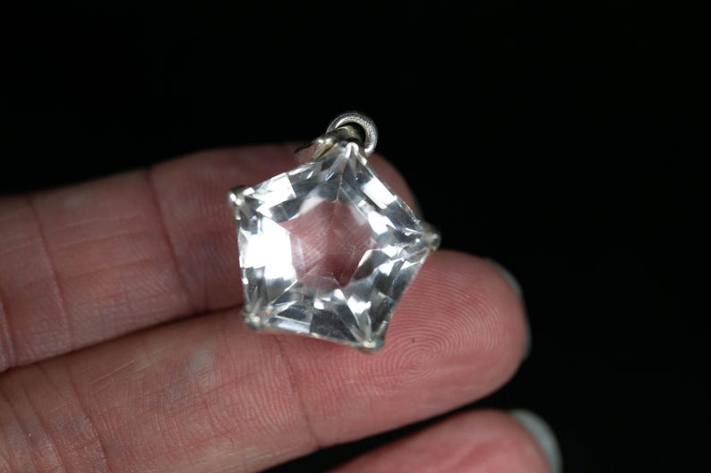 May include: A clear, faceted, star-shaped pendant made of a transparent material. The pendant features a metallic silver-toned setting along its edges and includes a small circular metal loop at the top for attachment to a chain or cord.
