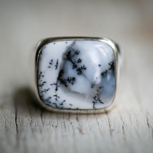 May include: A square-faced ring with a white stone featuring black dendritic inclusions. The ring has a silver-colored metal band. The stone's pattern resembles a delicate, black tree branch design.
