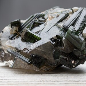 May include: A close-up of a mineral specimen featuring dark green tourmaline crystals embedded in a matrix of quartz. The crystals are elongated and have a glassy appearance, with a dark, almost black colour at the tips.