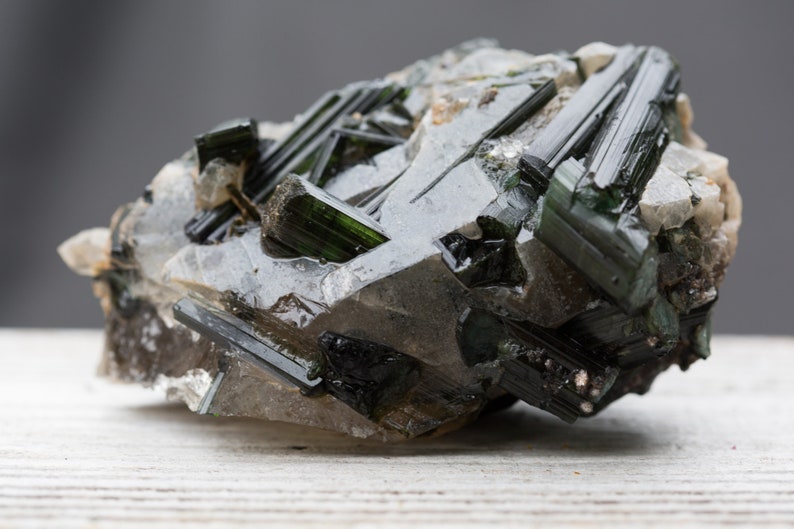 May include: A close-up of a mineral specimen featuring dark green tourmaline crystals embedded in a matrix of quartz. The crystals are elongated and have a glassy appearance, with a dark, almost black colour at the tips.