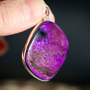 May include: A vibrant purple pendant with a silver-colored bezel. The gemstone has a polished, smooth surface with dark purple and black marbling. The pendant is held by a hand against a dark background. Jewelry.