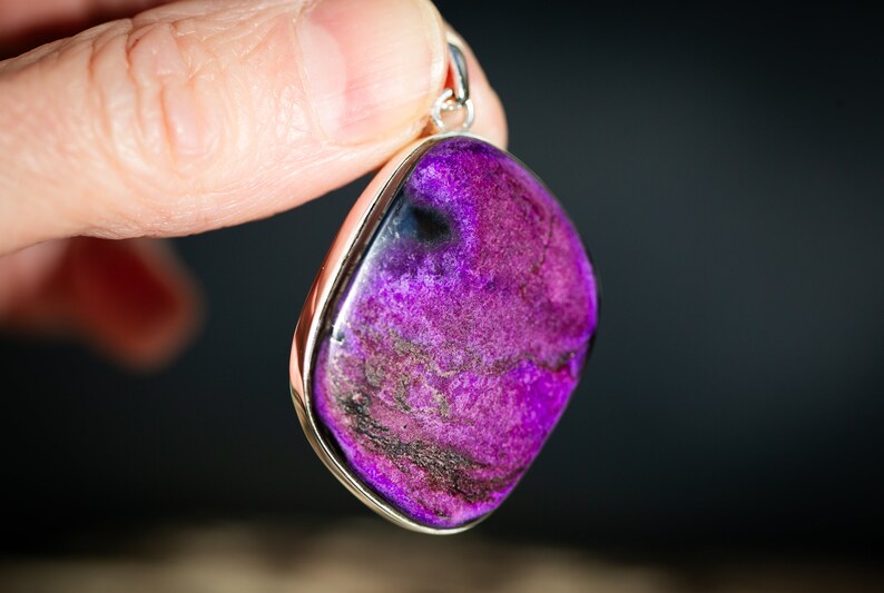 May include: A vibrant purple pendant with a silver-colored bezel. The gemstone has a polished, smooth surface with dark purple and black marbling. The pendant is held by a hand against a dark background. Jewelry.