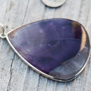 May include: A teardrop-shaped pendant with a polished, deep purple amethyst stone set in a silver bezel. The pendant has a small silver loop at the top for a chain. The stone's surface has natural variations.
