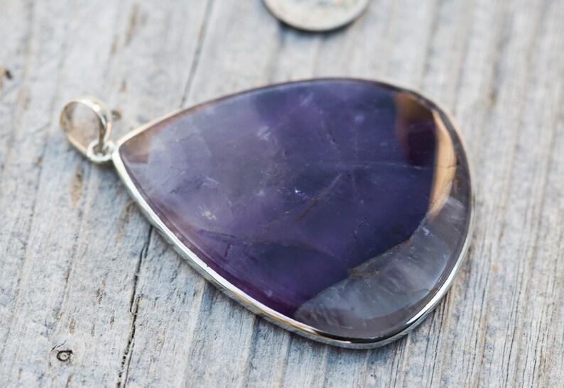 May include: A teardrop-shaped pendant with a polished, deep purple amethyst stone set in a silver bezel. The pendant has a small silver loop at the top for a chain. The stone's surface has natural variations.