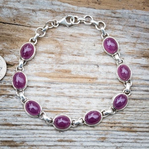 May include: A silver bracelet with a chain link design and oval-shaped purple gemstones. The bracelet is shown on a wooden surface.