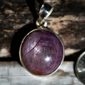 May include: A round, purple pendant with a silver bail. The pendant has a polished, gemstone appearance with a silver bezel. The gemstone has a unique pattern. The pendant is likely for a necklace.